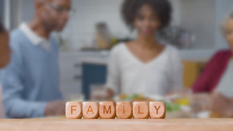 Concept-With-Wooden-Letter-Cubes-Or-Dice-Spelling-Family-Against-Background-Of-People-Eating-Meal-At-Home